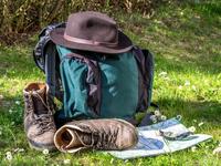Bild vergrößern: Ein Rucksack steht mit Hut, Wanderkarte und Kompass am Wegesrand. Neben ihm liegt ein Paar Wanderschuhe.